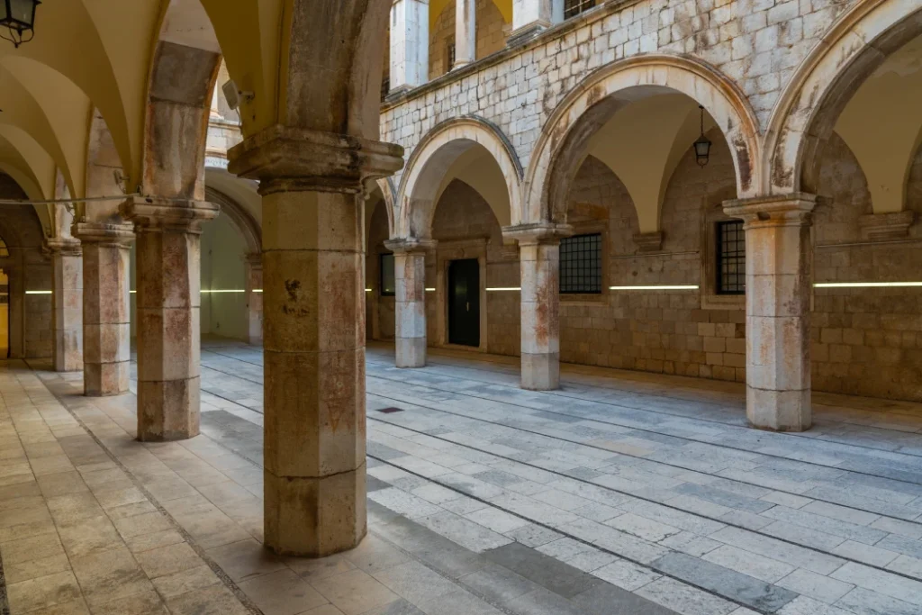 Interior view of Sponza Palace in Dubrovnik featuring stone columns, arches, and patterned flooring, with natural light illuminating the historical architecture.