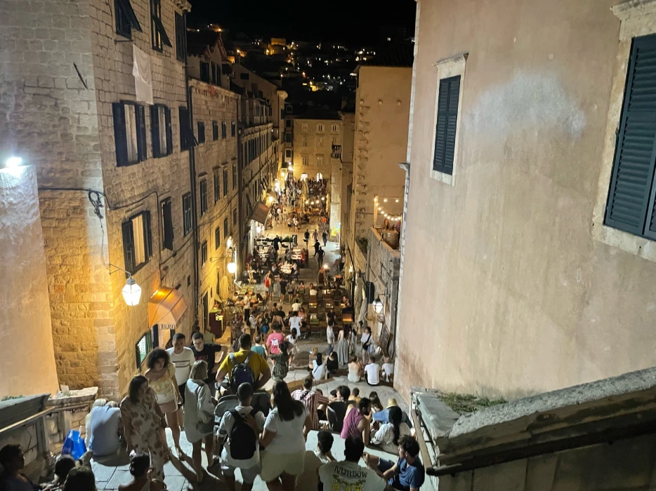A bustling evening scene at the Jesuit Stairs in Dubrovnik, with people descending the famous stone steps amidst historic buildings. The warm glow of street lights illuminates the vibrant nightlife and rich architectural heritage.