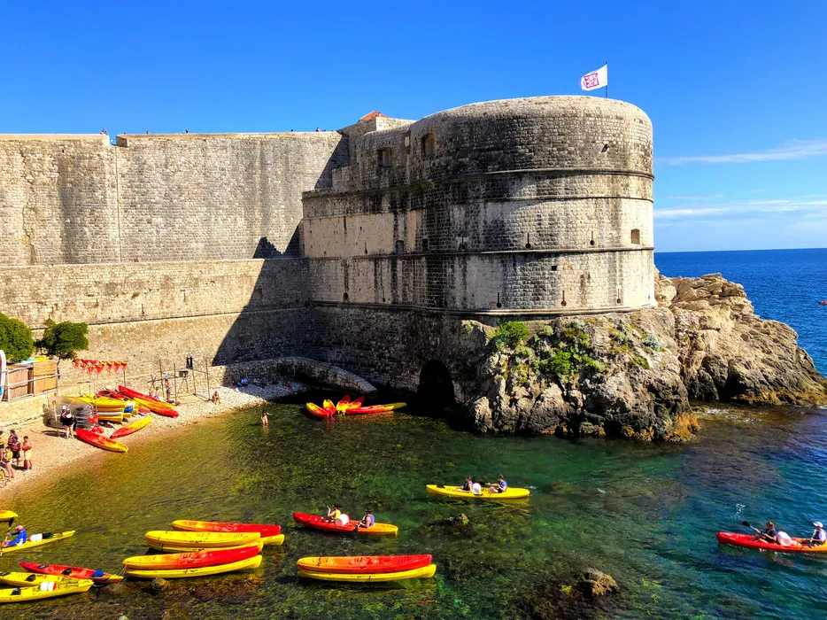 Kayakers enjoying the clear waters near the base of Dubrovnik's Pile Harbour, with the city's ancient fortress walls towering overhead under a clear blue sky.