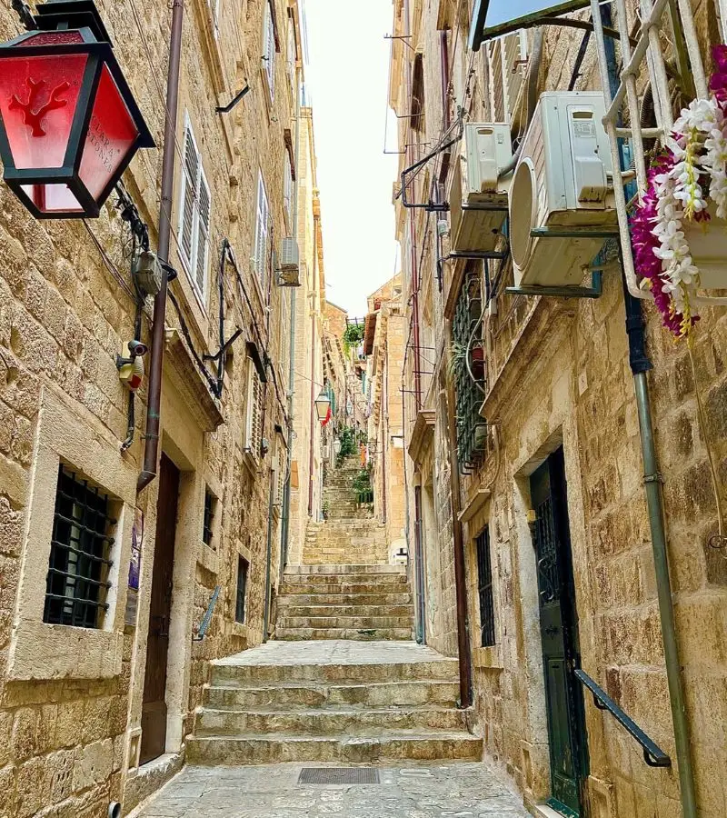 Narrow stone-paved street ascending between aged limestone buildings in Old Town Dubrovnik, with air conditioning units on walls and a red lantern hanging to the left.