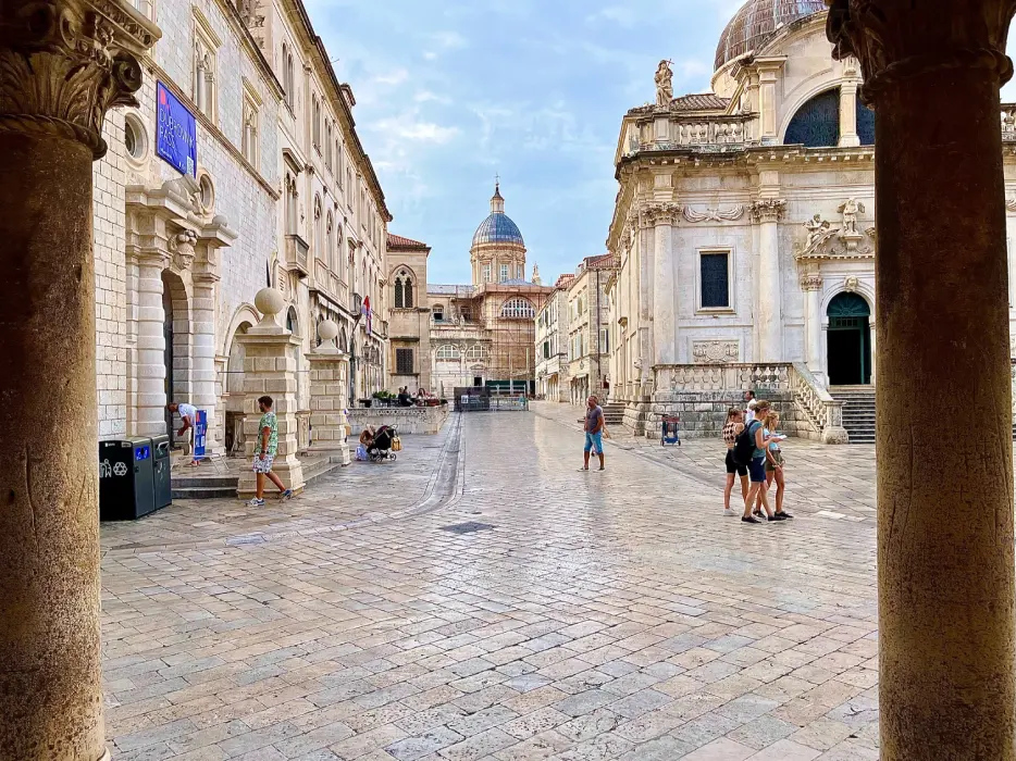 A serene day outside the Sponza Palace in Dubrovnik, with tourists meandering around the historical square framed by classical architecture and a cobblestone pavement gleaming under the soft sunlight.