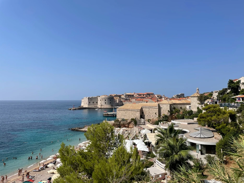 Panoramic view of Banje Beach in Dubrovnik, with the historic city walls and clear blue waters of the Adriatic Sea, as beachgoers enjoy the sun and sea.