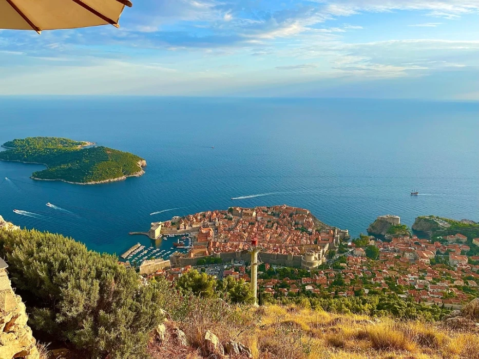 A panoramic view from a high vantage point overlooking the old town of Dubrovnik, Croatia, with the Adriatic Sea in the background. A lush green island and boats can be seen on the water, while terracotta rooftops dominate the town's landscape