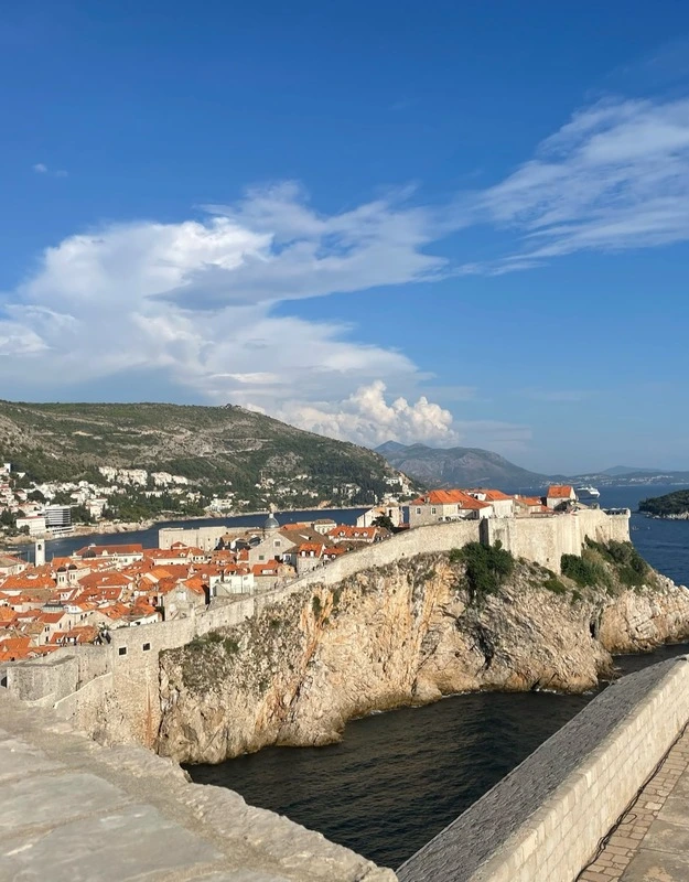 Panoramic view from Fort Lovrijenac showcasing the historic Old Town of Dubrovnik, with its iconic orange rooftops, surrounded by the Adriatic Sea and framed by a blue sky with wispy clouds.