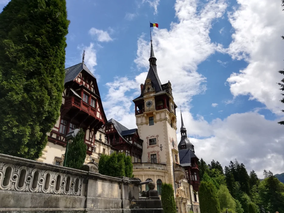 View of the historical Peles Castle in Romania, displaying its charming wooden details and spired towers, under a blue sky with fluffy clouds, flanked by green trees.