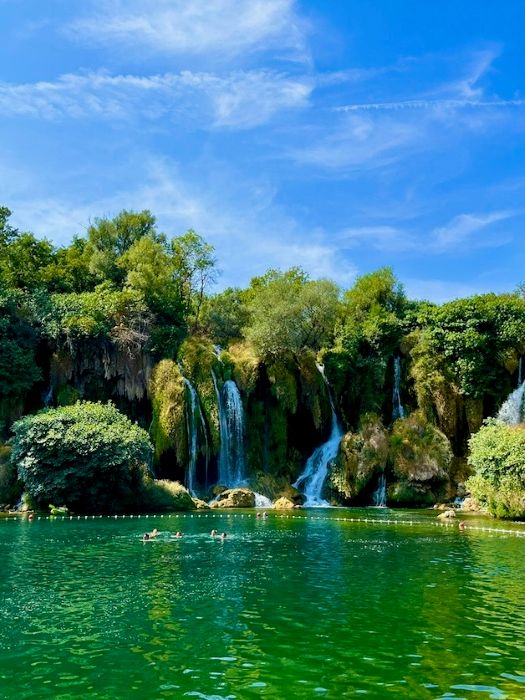 People swimming in the serene waters of the Kravica Waterfalls basin with surrounding cascades.