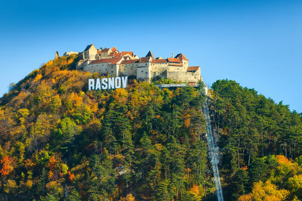 The historic Rasnov Citadel perched on a hilltop surrounded by autumn-colored trees with the bold white letters 'RASNOV' in the foreground, reminiscent of the Hollywood sign, under a clear blue sky.