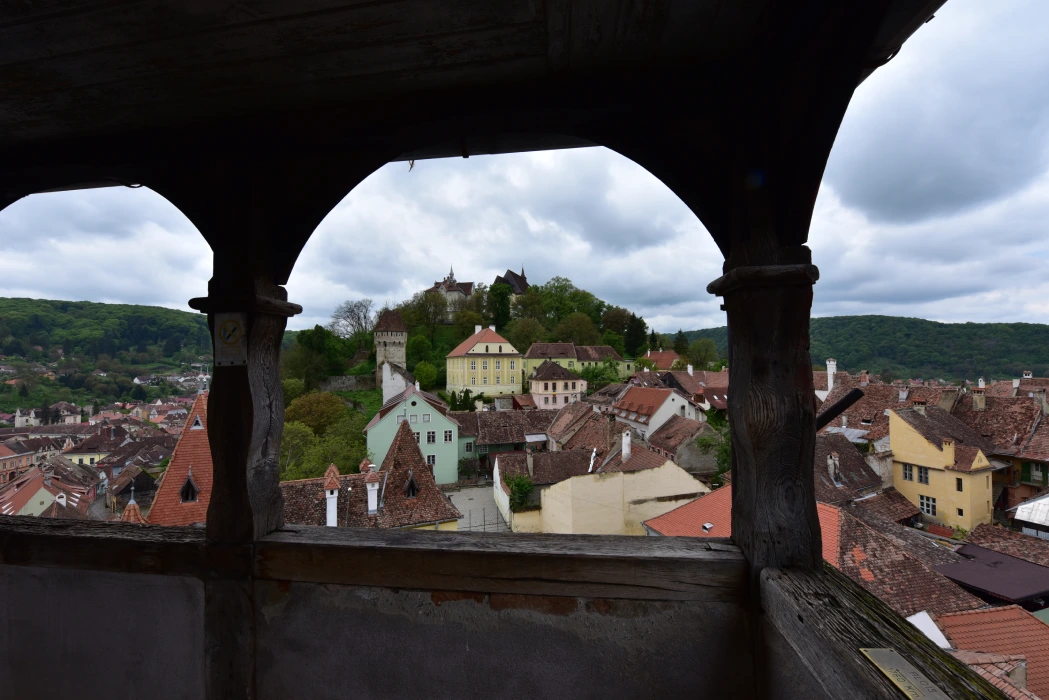 A scenic view of the historical town of Sighisoara, Romania, captured from the balcony of its iconic Clock Tower. The balcony's carved wooden arches frame the rolling hills in the background and the colorful, tiled roofs of the medieval houses below, under a cloudy sky.