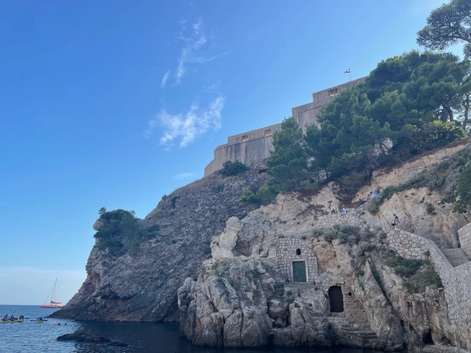 View of Fort Lovrijenac perched atop a rocky cliff in Dubrovnik, with people ascending the stone stairway against a backdrop of blue skies and a sailing boat on the water.