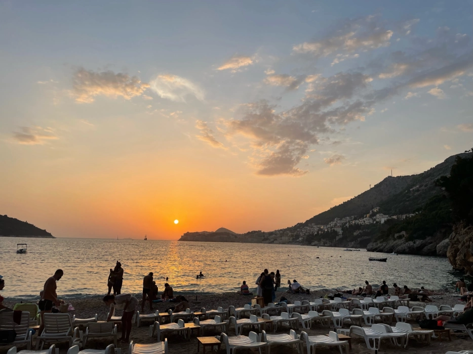 Sunset at Sveti Jakov Beach, Dubrovnik, with the sun dipping below the horizon, silhouetting beachgoers and boats against an orange sky, while shadows stretch across the sandy shore lined with lounge chairs.