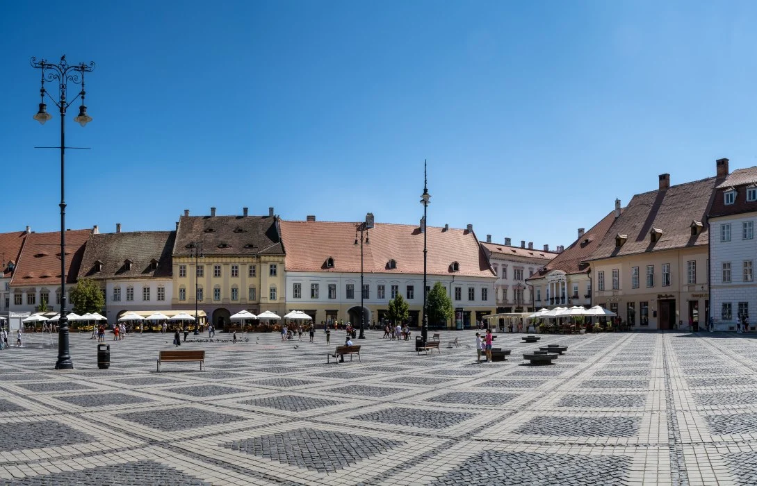 A sunny day at The Large Square in Sibiu showcasing the patterned cobblestone, colorful two-story buildings with terracotta roofs, street lamps, and a scattering of people and outdoor cafes under white umbrellas