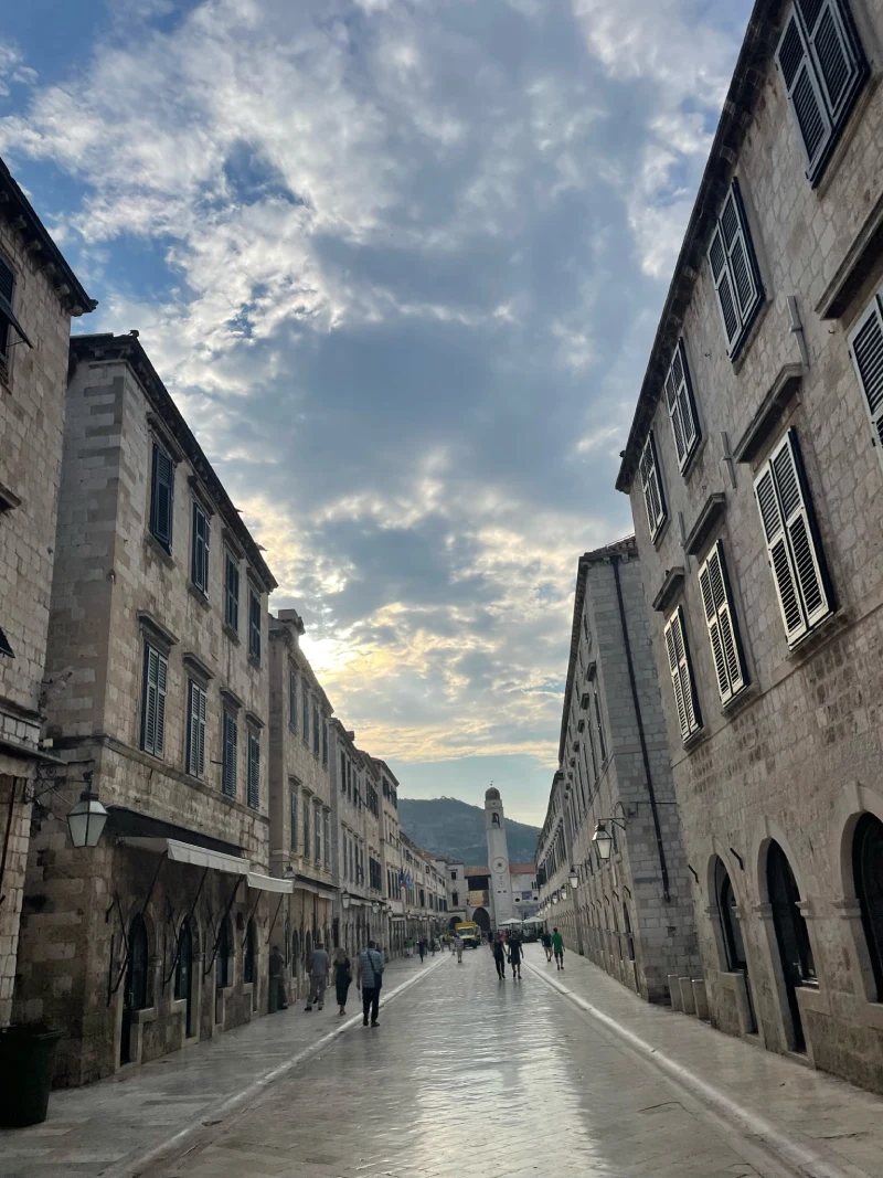 Pedestrians stroll down the Stradun, Dubrovnik's main limestone-paved thoroughfare, flanked by buildings and shops, under a partially cloudy sky with the sun peeking through, creating a serene early evening ambiance.