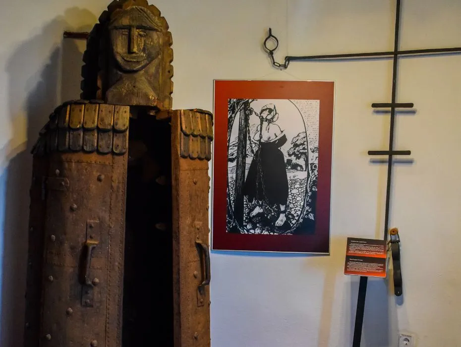A medieval wooden torture mask stands open next to a wall displaying an illustration of a woman and several iron cross-shaped wall fixtures, part of the torture exhibition at Bran Castle