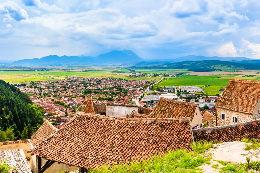 Panoramic view of a picturesque town from the Rasnov Citadel, showcasing terracotta rooftops, dense housing, and the expansive green plains stretching to distant mountains under a partly cloudy sky.