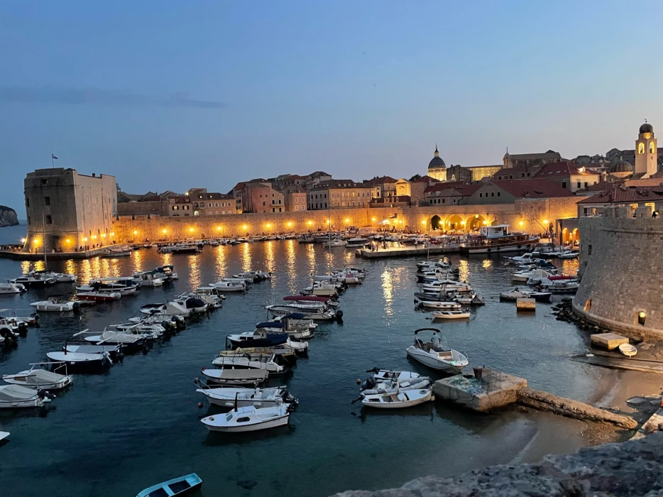 The picturesque Old Harbour of Dubrovnik, captured from the Ploče Gate during twilight. The historical fortress walls are illuminated, bordering the calm Adriatic Sea which hosts an array of boats and yachts, while the warm glow of street lights reflects gently on the water's surface.