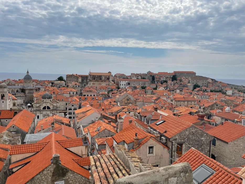 An elevated view of Dubrovnik's Old Town, showcasing the dense array of terracotta rooftops, various church domes and bell towers, under a sky dotted with fluffy clouds. The historic architecture spreads towards the Adriatic Sea, highlighting the city's well-preserved medieval charm