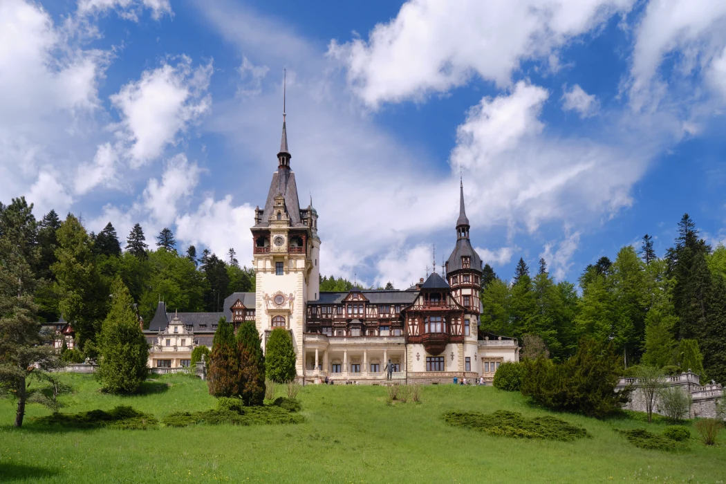 The picturesque Peleș Castle in Romania, showcasing its ornate towers and intricate woodwork against a backdrop of lush greenery and a blue sky speckled with clouds.