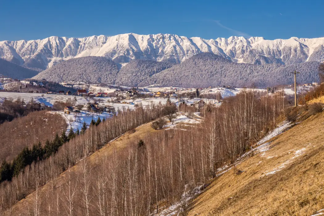 A scenic winter view of Piatra Craiului Mountains in Romania, showcasing a village nestled in a valley with patchy snow cover, barren trees, and the majestic snow-capped mountain range in the background under a clear blue sky.