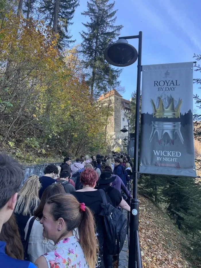 Crowd of visitors queueing along a narrow path leading up to Bran Castle on a sunny day, with a banner reading 'ROYAL BY DAY WICKED BY NIGHT' featuring an image of a crown and bat wings, hinting at a Halloween event at the castle.
