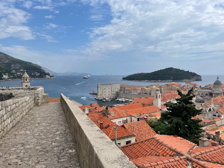 A picturesque view of Dubrovnik's Old Town from the city walls, showcasing terracotta rooftops, the Adriatic Sea, and the historic city walls under a partly cloudy sky. A cruise ship and smaller boats can be seen near the harbour.