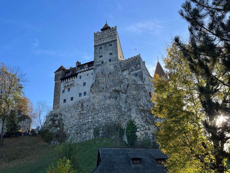 Bran Castle perched atop a rocky cliff surrounded by lush greenery, with the sun casting a soft glow through the trees, in a serene autumn setting