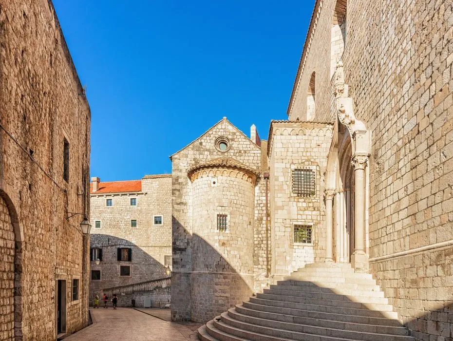 A sunlit view of the exterior of the Dominican Monastery in Dubrovnik, showcasing its medieval architecture. The scene features robust stone walls, a prominent staircase leading to a sculpted doorway, and a round tower with arched windows, under a clear blue sky.