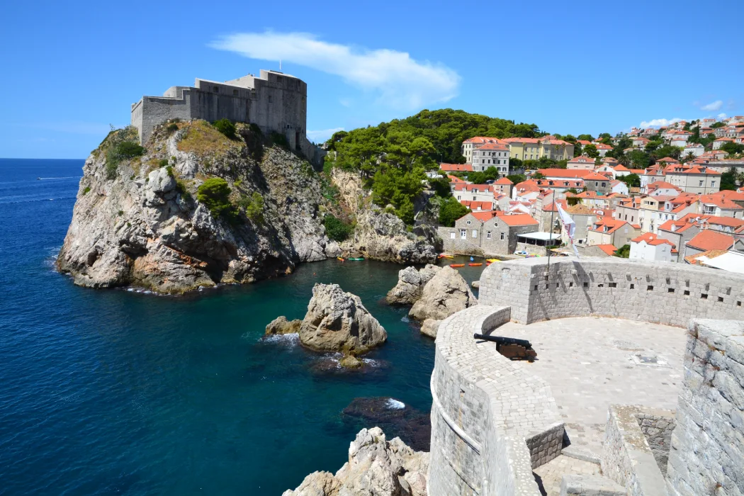 View of Fort Bokar in Dubrovnik, perched on a rugged cliff overlooking the Adriatic Sea. The fortress is surrounded by clear blue waters and jagged rocks, with a backdrop of the historic city featuring terracotta-roofed buildings and lush greenery.