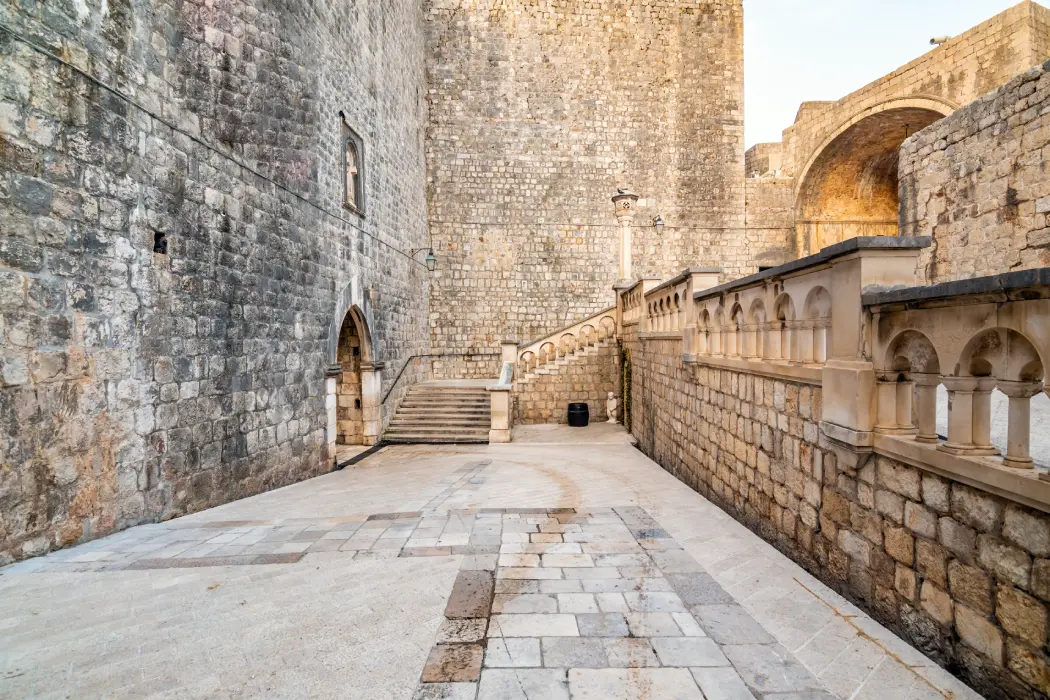 Empty courtyard at Pile Gate, Dubrovnik, captured in the soft light of early morning. The image features ancient stone walls and archways, a set of steps leading to a wooden door, and elegant balustrades.
