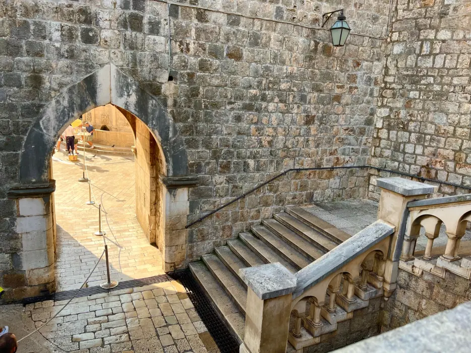 Stone archway entrance at Pile Gate, leading into the old town of Dubrovnik. The view captures the historical texture of the weathered stone walls, a stone staircase, and balustrades, under the warm sunlight, with tourists visible in the distance exploring the site.