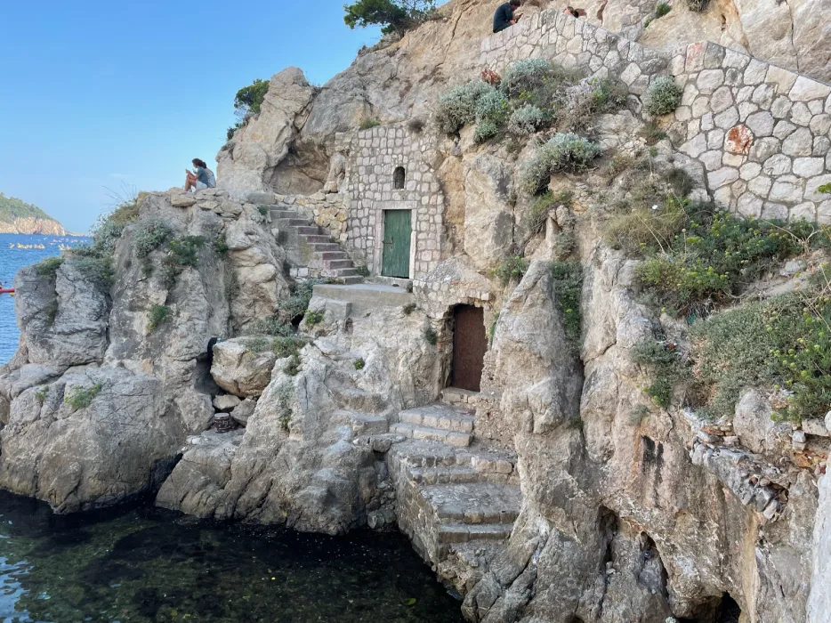 Rustic stone structures built into a rocky cliffside beside the sea at Dubrovnik's West Harbour. Stone stairs lead up to a small door and seating area where a couple sits enjoying the view, with clear blue waters below