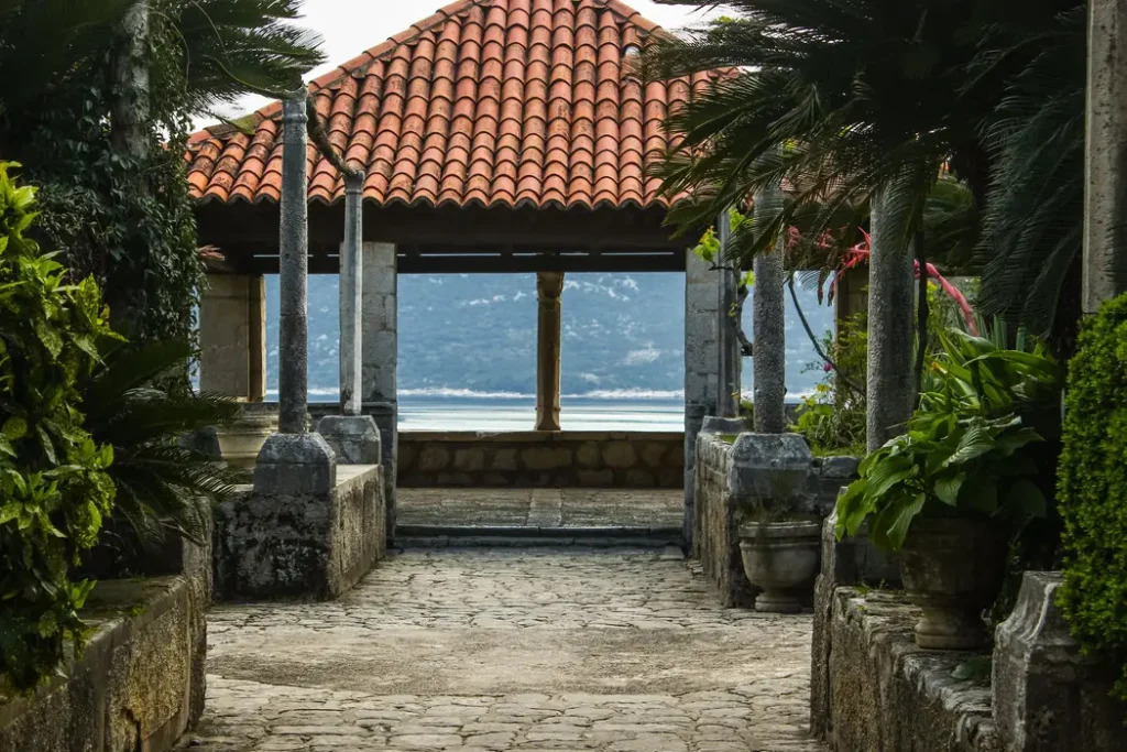 The Trsteno Arboretum Belvedere Pavilion in Croatia, showcasing a stone pathway leading to the historic open-air structure with a red tile roof, surrounded by verdant tropical plants, with the tranquil Adriatic Sea and distant mountains providing a picturesque backdrop.