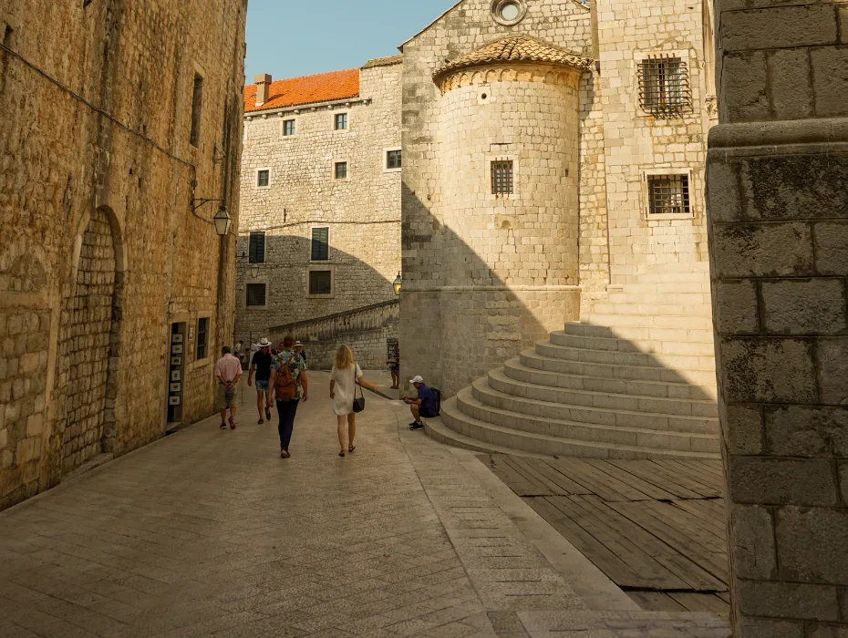 A bustling scene on St. Dominic Street in Dubrovnik with tourists walking along a cobblestone pathway, flanked by ancient stone buildings