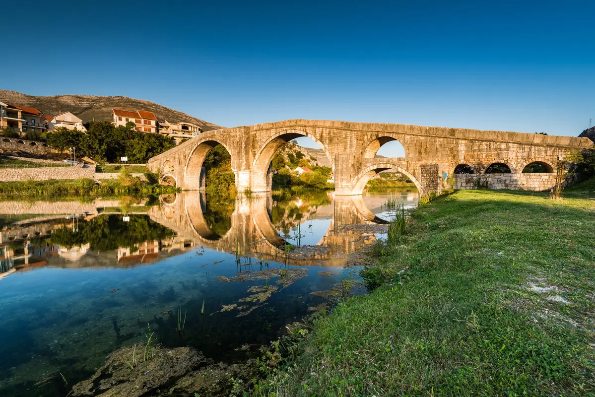 The Arslanagić Bridge in Trebinje, Bosnia and Herzegovina, featuring multiple arches reflected in the river below. The surrounding area includes greenery and residential buildings, all set against a clear blue sky.