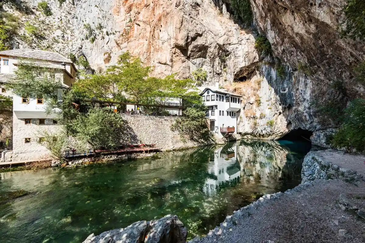 The Blagaj Tekke, a Dervish monastery situated at the base of a cliff beside the Buna River in Blagaj, Bosnia and Herzegovina. The setting features clear, reflective water, rocky cliffs, and lush greenery surrounding the white stone buildings of the monastery.