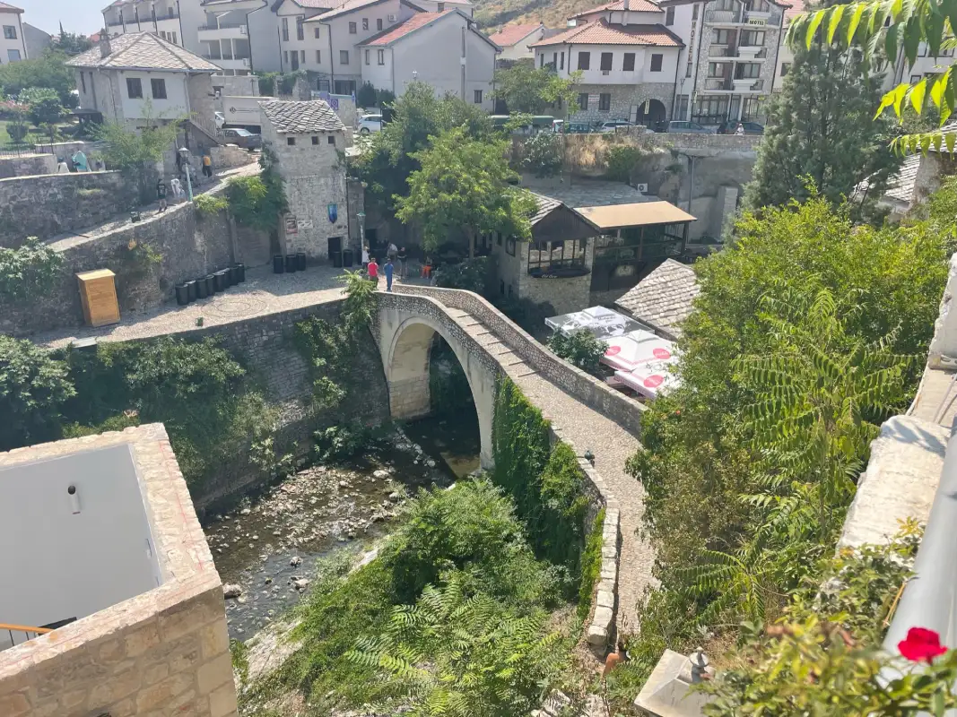 The Crooked Bridge (Kriva Ćuprija) in Mostar, Bosnia and Herzegovina. The stone bridge arches over a small, rocky stream, surrounded by greenery and buildings. Nearby, people can be seen exploring the area.