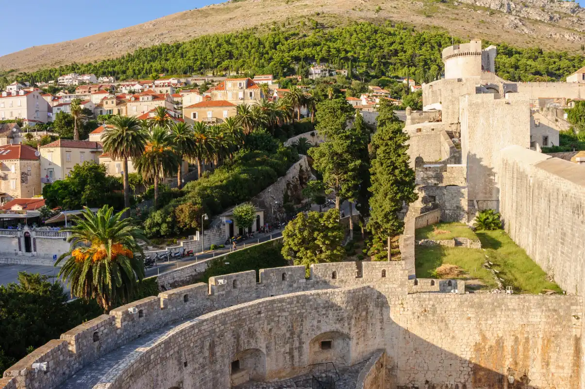 A view of the Dubrovnik City Walls on a sunny day. The image showcases its aged stone walls and round defensive towers, surrounded by greenery and rows of orange-roofed houses, with a backdrop of a hilly landscape