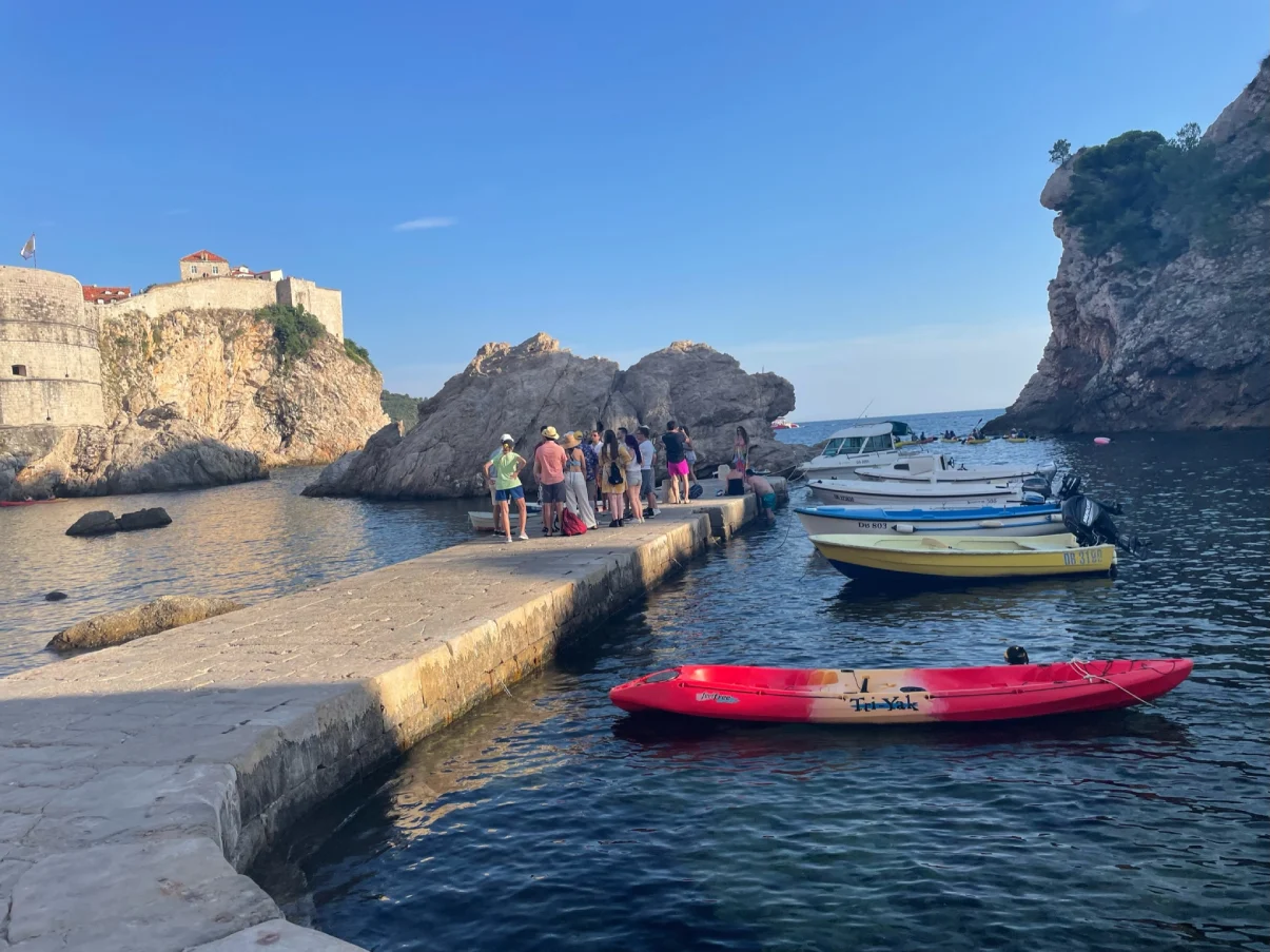 Tourists on a Game of Thrones-themed tour walking along a narrow stone jetty at Dubrovnik's West Pier (Blackwater Bay). The setting includes blue waters, moored boats including a red kayak, and rugged cliffs.