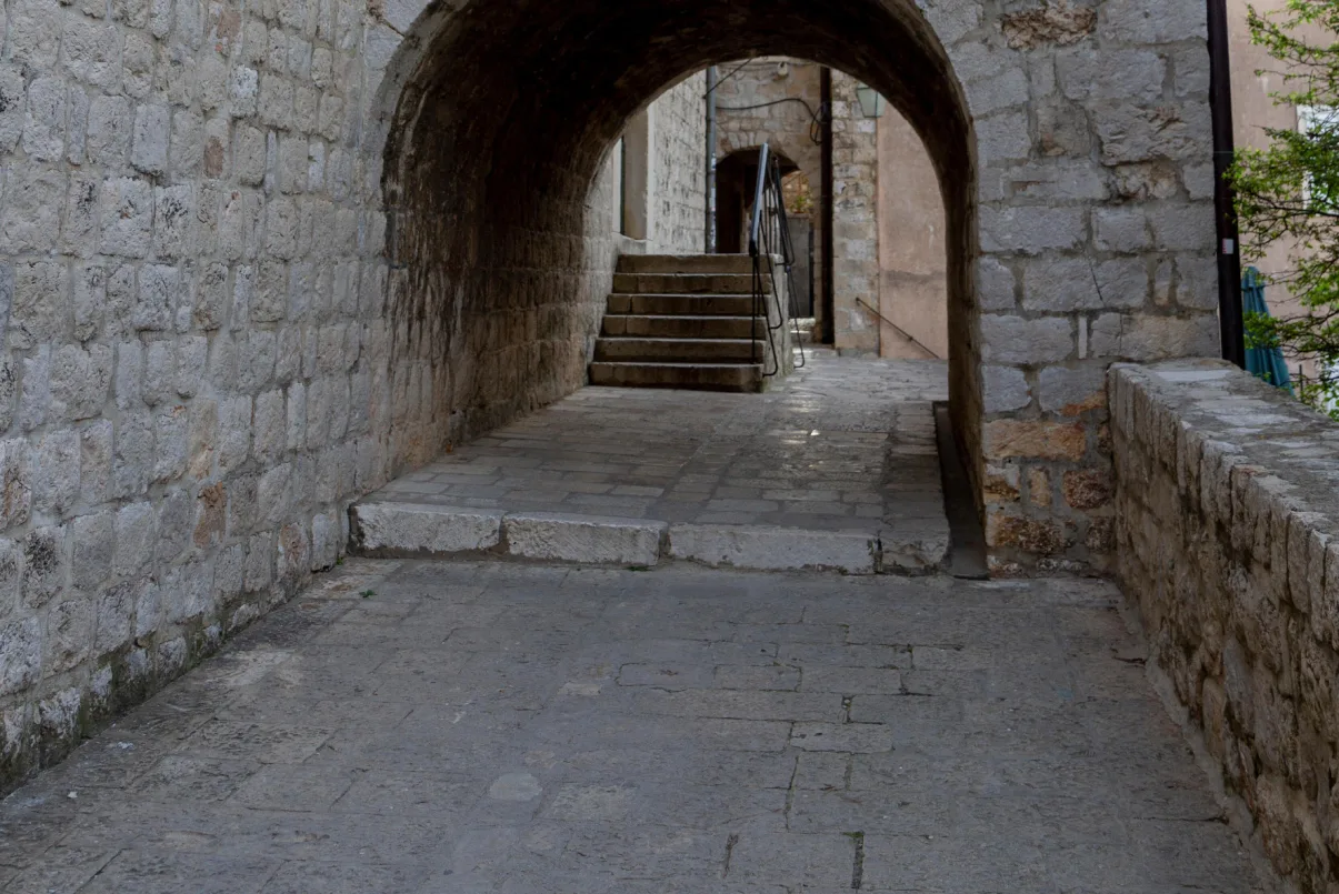 Stone archway at the Ethnographic Museum in Dubrovnik, which served as a filming location for Game of Thrones. The pathway leads through the arch to a set of stairs, with textured stonework typical of medieval European architecture. 