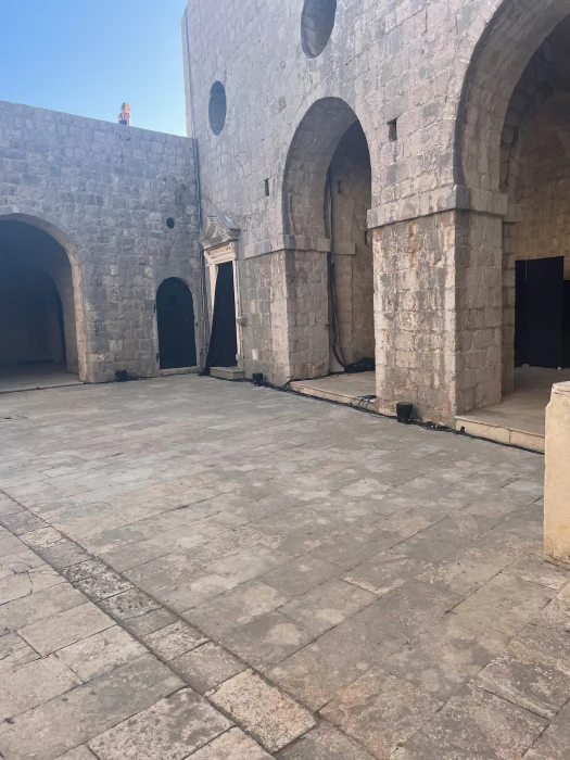 This image showcases another angle of Fort Lovrijenac's courtyard in Dubrovnik, Croatia. It highlights the arched stone architecture, smooth stone floor, and circular wall openings under a clear blue sky. This area is famous for being featured in Game of Thrones.