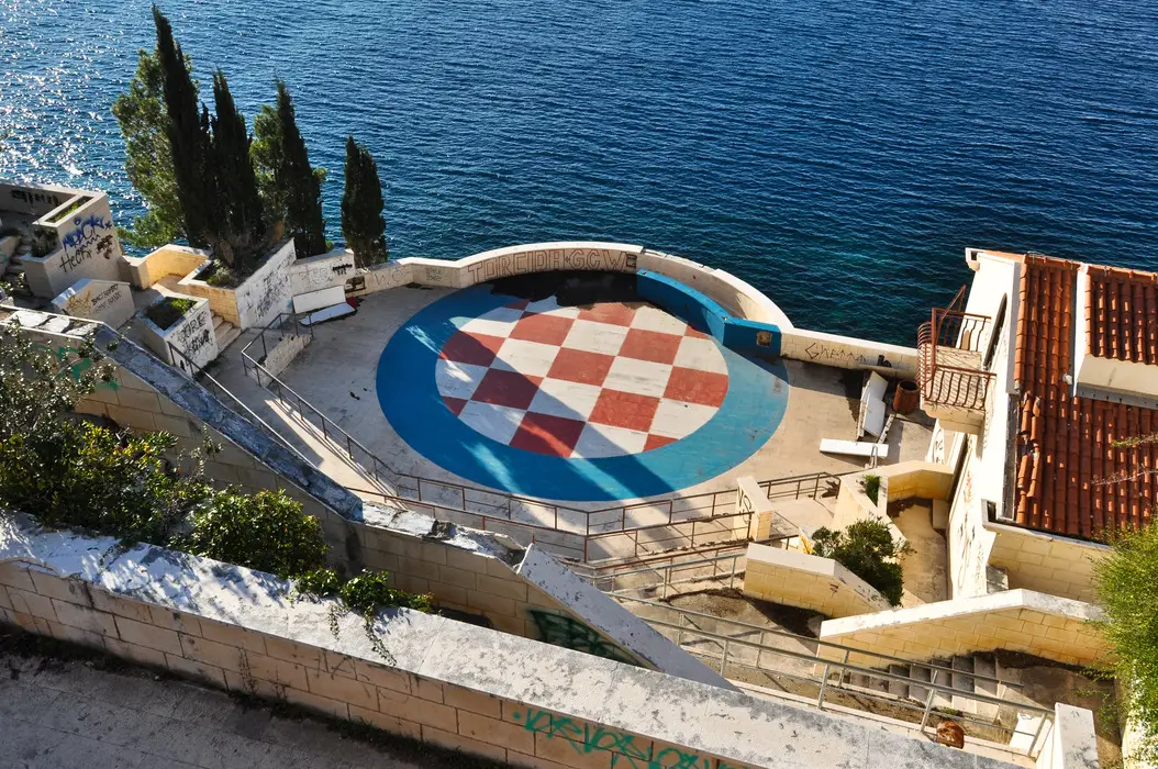 Overhead view of the abandoned Hotel Belvedere amphitheater near Dubrovnik, Croatia, overlooking the Adriatic Sea. The circular stage features a large, colorful checkerboard pattern, surrounded by graffiti-covered concrete seating, amidst overgrown vegetation and adjacent to terracotta-roofed structures.