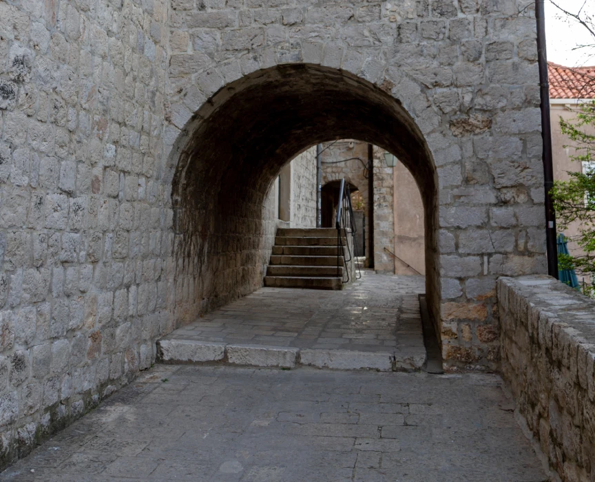 Stone archway at the Ethnographic Museum in Dubrovnik, which also served as a filming location for Game of Thrones. The pathway leads through the arch to a set of stairs, with historic, textured stonework typical of medieval European architecture. Light filters through the arch, highlighting the old-world charm of the location.