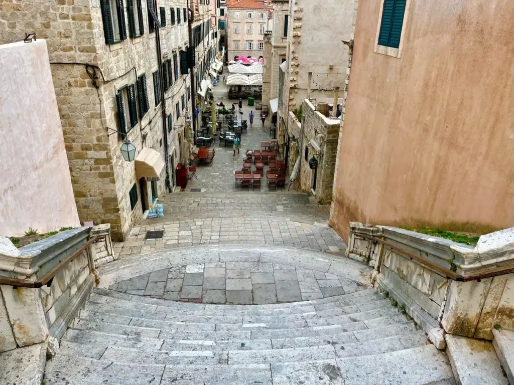 The image shows Jesuit Stairs in Dubrovnik, Croatia, a famous filming location for Game of Thrones. The stone steps descend into a narrow street lined with buildings and outdoor dining tables.