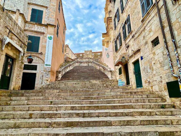 The iconic Jesuit Stairs in Dubrovnik, Croatia, made famous as a filming location in Game of Thrones. The wide stone staircase is surrounded by buildings with shuttered windows and weathered walls, leading up to the Baroque-style Church of St. Ignatius. The sky above is bright and partly cloudy.