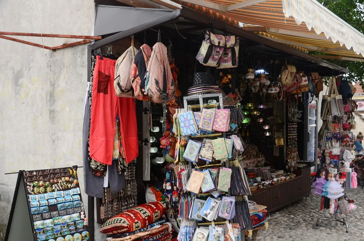 A market stall in the Old Bazaar of Mostar, Bosnia and Herzegovina, displaying an array of colourful goods. Hanging bags, bright red trousers, intricately designed wallets, and various souvenirs are prominently featured.