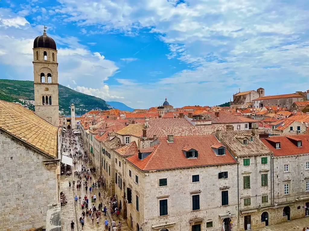 This image showcases the Old Town of Dubrovnik, Croatia, as seen from the City Walls. The view highlights terracotta rooftops, stone buildings, and the bell towers under a partly cloudy blue sky. The famous street 'The Stradun' is bustling with tourists below.  