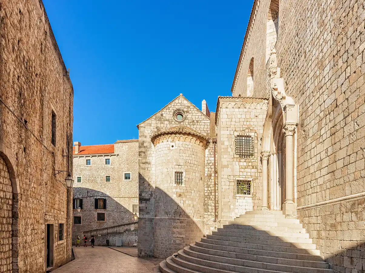 A sunlit view of the exterior of the Dominican Monastery in Dubrovnik. The scene features robust stone walls, a prominent staircase leading to a sculpted doorway, and a round tower with arched windows, under a clear blue sky.