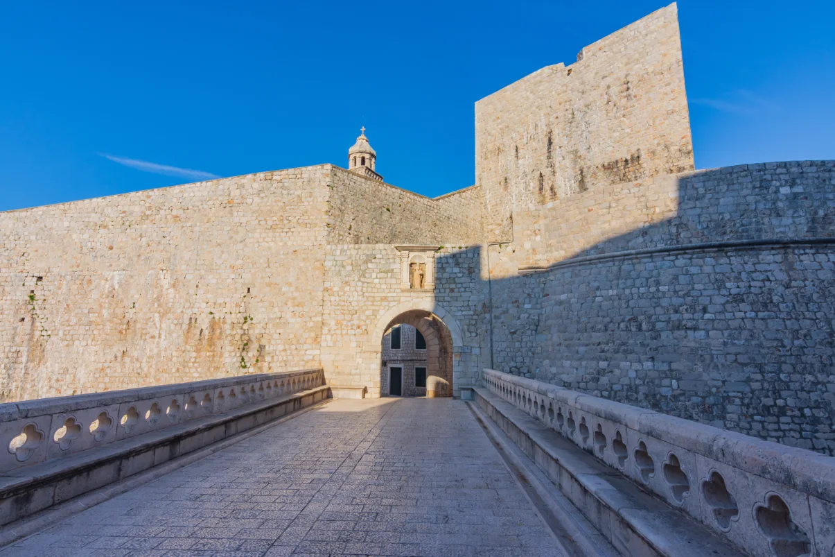 View of Ploce Gate, a historic stone entrance to Dubrovnik's old town, under a clear blue sky. The gate features a stone bridge leading to a massive archway, flanked by robust medieval walls and a small domed structure on top