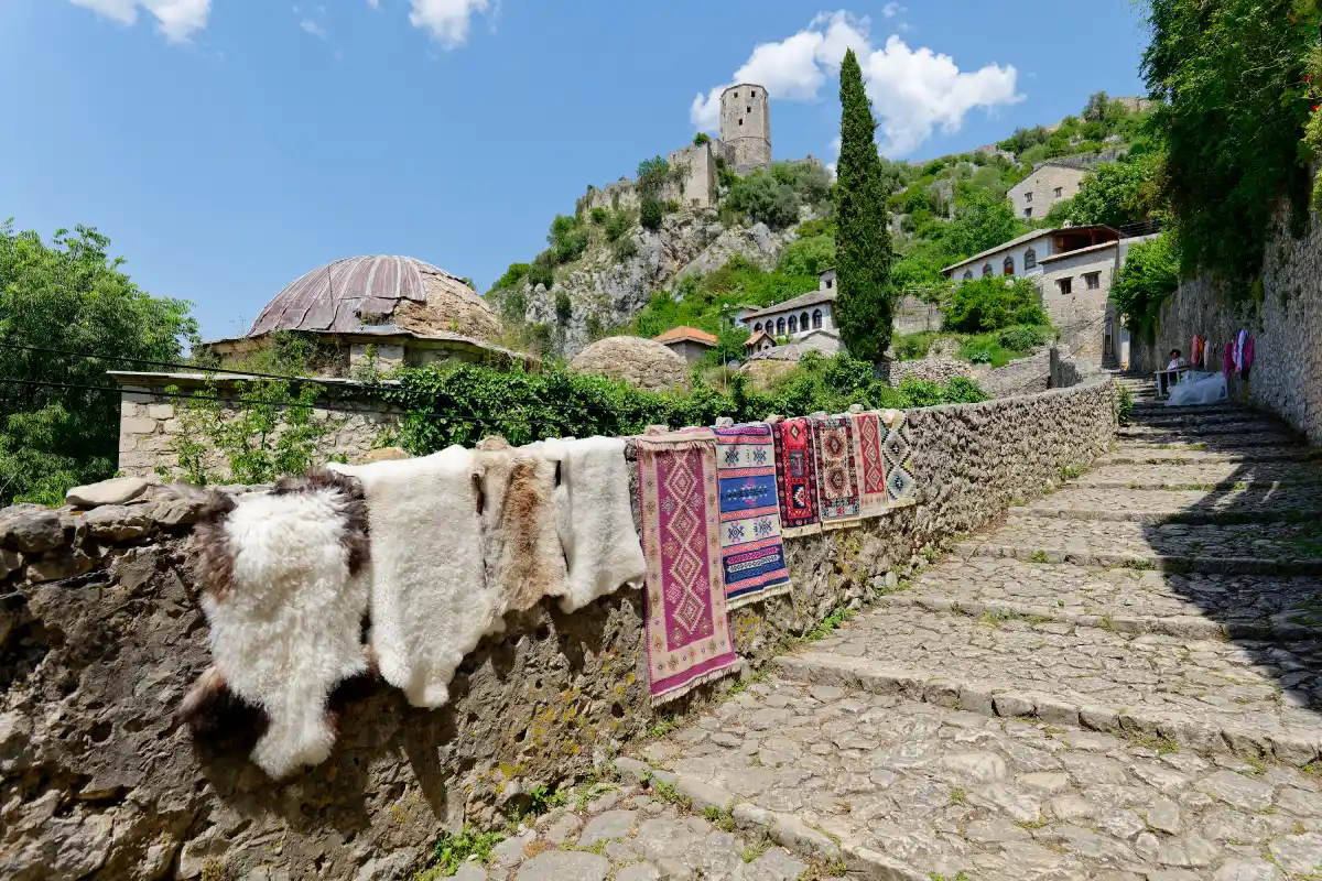 A view of Počitelj, a village in Bosnia and Herzegovina. The stone pathway is lined with traditional rugs and sheepskins displayed on a stone wall, leading up to ancient stone buildings and a round tower on a hill, surrounded by greenery under a bright blue sky.