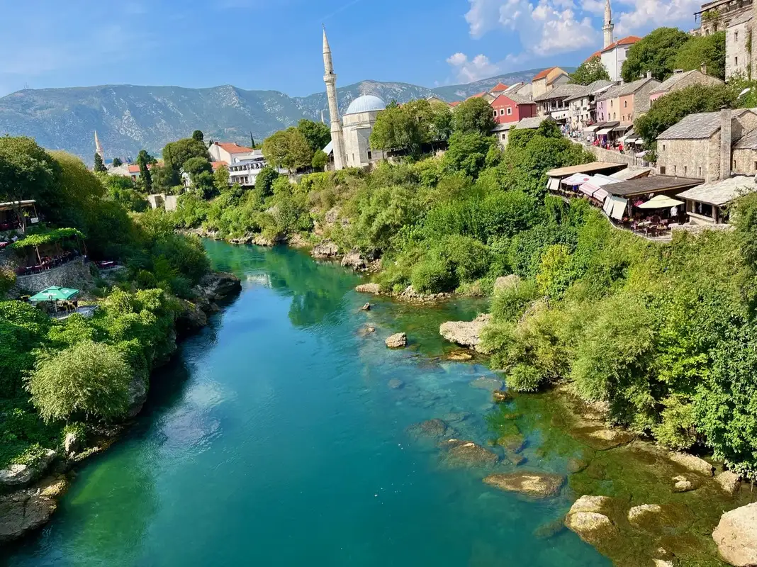 A view of Mostar, Bosnia and Herzegovina, from the Stari Most bridge. The Neretva River flows through the lush green landscape, with traditional buildings and a mosque visible on the right side, set against a backdrop of mountains. The clear blue sky is above adding to the tranquil atmosphere of the scene.