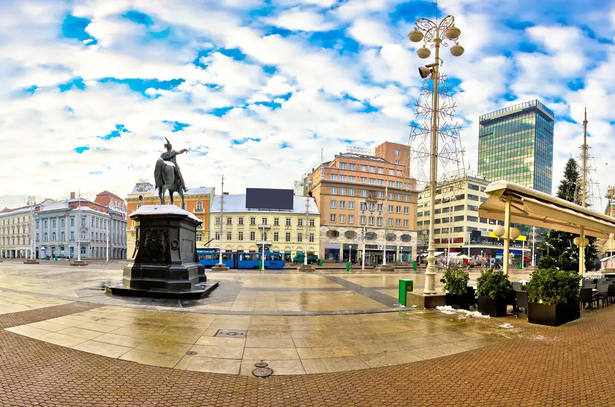 Ban Jelačić Square in Zagreb's Lower Town that shows the statue of Ban Josip Jelačić on horseback at the centre. Surrounding the square are a variety of historical and modern buildings under a bright, partly cloudy sky